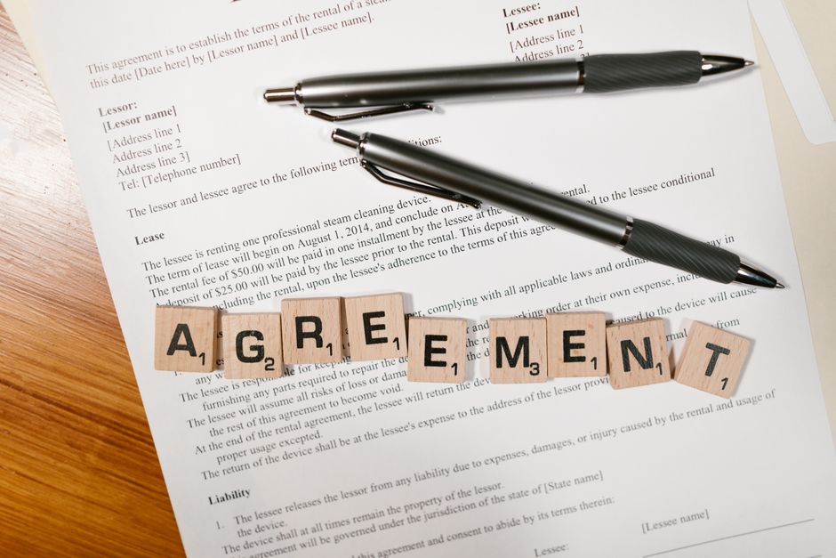 A document with Scrabble tiles spelling ’AGREEMENT’ and two pens, symbolizing contract signing.